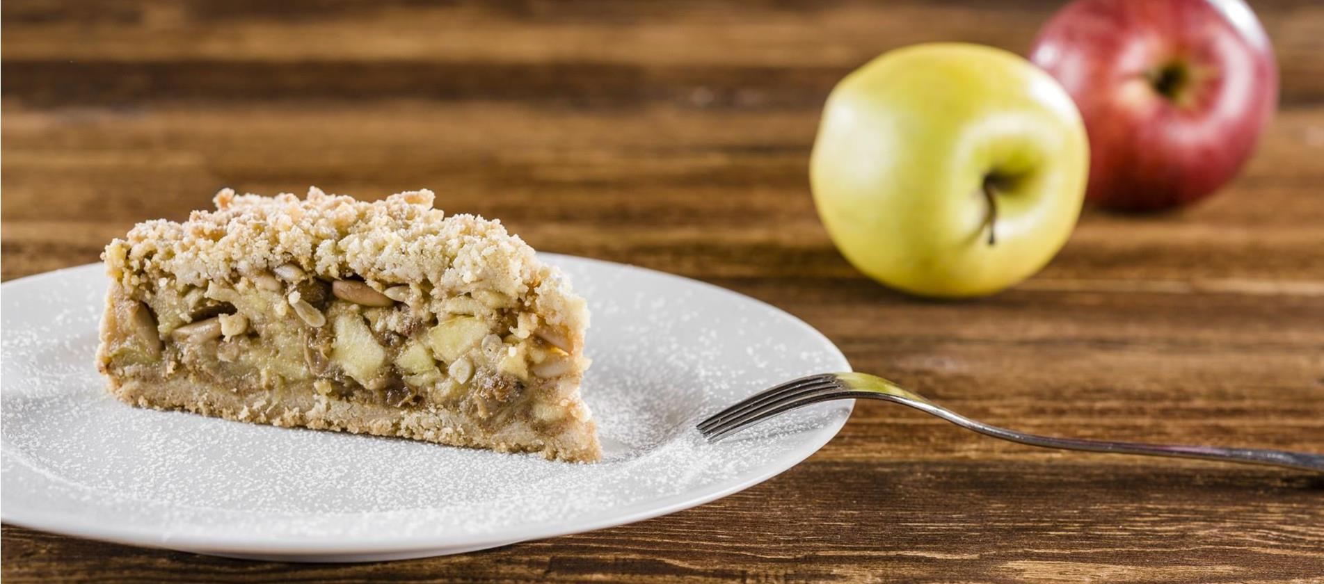 Ein Stück Apfelstreuselkuchen auf einem weißen Teller, bestäubt mit Puderzucker; im Hintergrund liegen zwei Äpfel, einer rot und einer gelb, auf einem Holztisch.