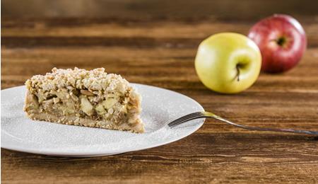 Ein Stück Apfelstreuselkuchen auf einem weißen Teller, bestäubt mit Puderzucker; im Hintergrund liegen zwei Äpfel, einer rot und einer gelb, auf einem Holztisch.
