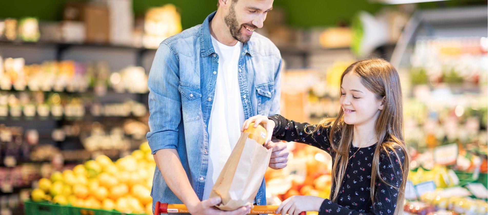 Ein Mann und ein Mädchen kaufen gemeinsam Äpfel in einem Supermarkt ein und legen sie in eine Papiertüte – im Hintergrund ist frisches Obst sichtbar.