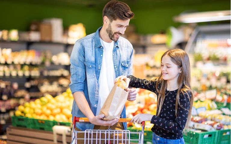 Ein Mann und ein Mädchen kaufen gemeinsam Äpfel in einem Supermarkt ein und legen sie in eine Papiertüte – im Hintergrund ist frisches Obst sichtbar.