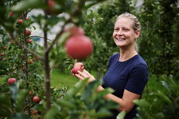 Apfelbäuerin Andrea Erlacher steht zwischen Apfelbäumen und hält mehrere rote Äpfel in den Händen.