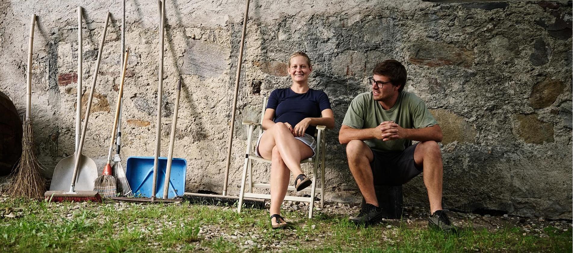 Andrea Erlacher und Jacob Gamper sitzen gemeinsam vor ihrem Hof neben verschiedenen Arbeitsgeräten.