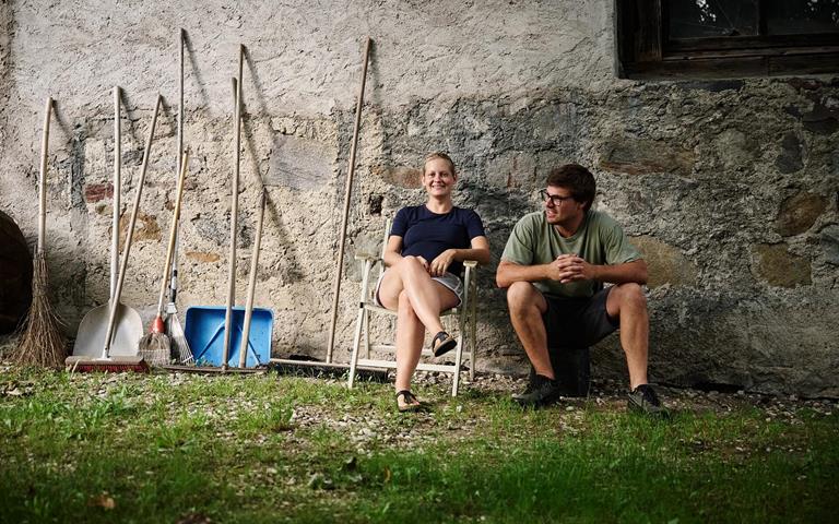 Andrea Erlacher und Jacob Gamper sitzen gemeinsam vor ihrem Hof neben verschiedenen Arbeitsgeräten.