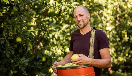 Apfelbauer steht in der Apfelwiese mit Kiste frischer Äpfel in der Hand und lächelt in die Kamera