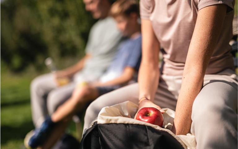 Person macht auf einer Bank Pause und greift zu einem roten Südtiroler Apfel g.g.A. aus dem Rucksack, während weitere Personen im Hintergrund entspannen.